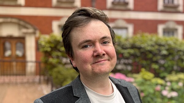 Portrait-style Liberal Democrat candidate announcement graphic featuring a young man standing outdoors in front of a red-brick building and garden with pink flowers. He is wearing a grey blazer over a light-coloured shirt and smiling slightly at the camera. The background is softly blurred. Overlaid text reads “Candidate Announcement”, “JAKE AUSTIN”, and “Makerfield”, with the Liberal Democrats logo at the bottom. Orange and dark blue gradient lighting effects frame the lower corners of the image.