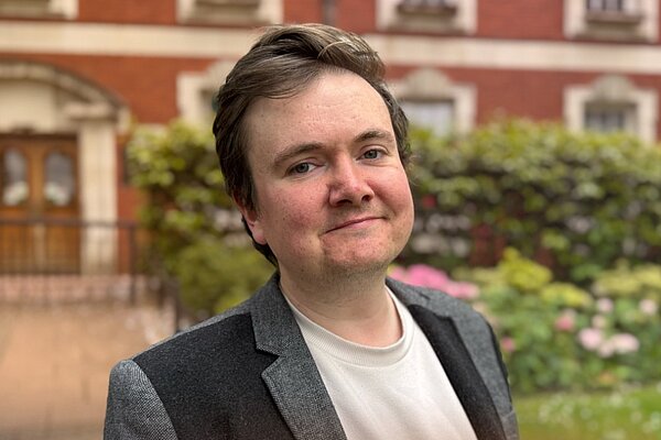 Portrait-style Liberal Democrat candidate announcement graphic featuring a young man standing outdoors in front of a red-brick building and garden with pink flowers. He is wearing a grey blazer over a light-coloured shirt and smiling slightly at the camera. The background is softly blurred. Overlaid text reads “Candidate Announcement”, “JAKE AUSTIN”, and “Makerfield”, with the Liberal Democrats logo at the bottom. Orange and dark blue gradient lighting effects frame the lower corners of the image.